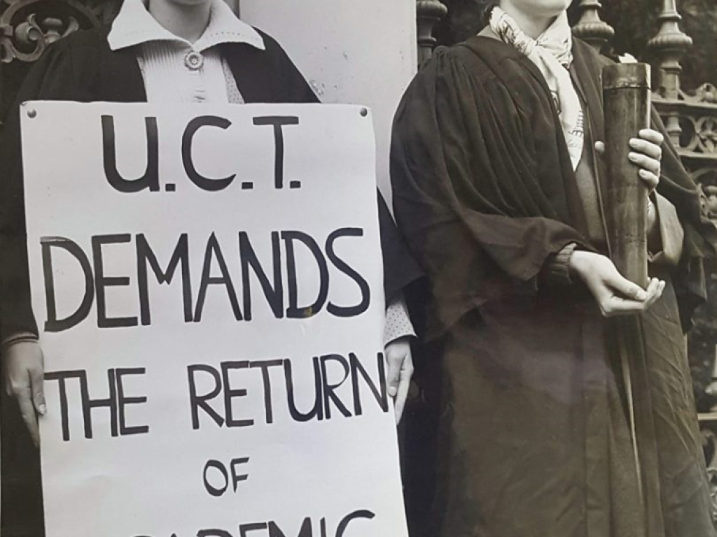 Women protesting in support of academic freedom. A woman holds a placard saying "UCT demands the return of academic freedom" beside a woman holding a lit torch. They are both wearing academic dress.